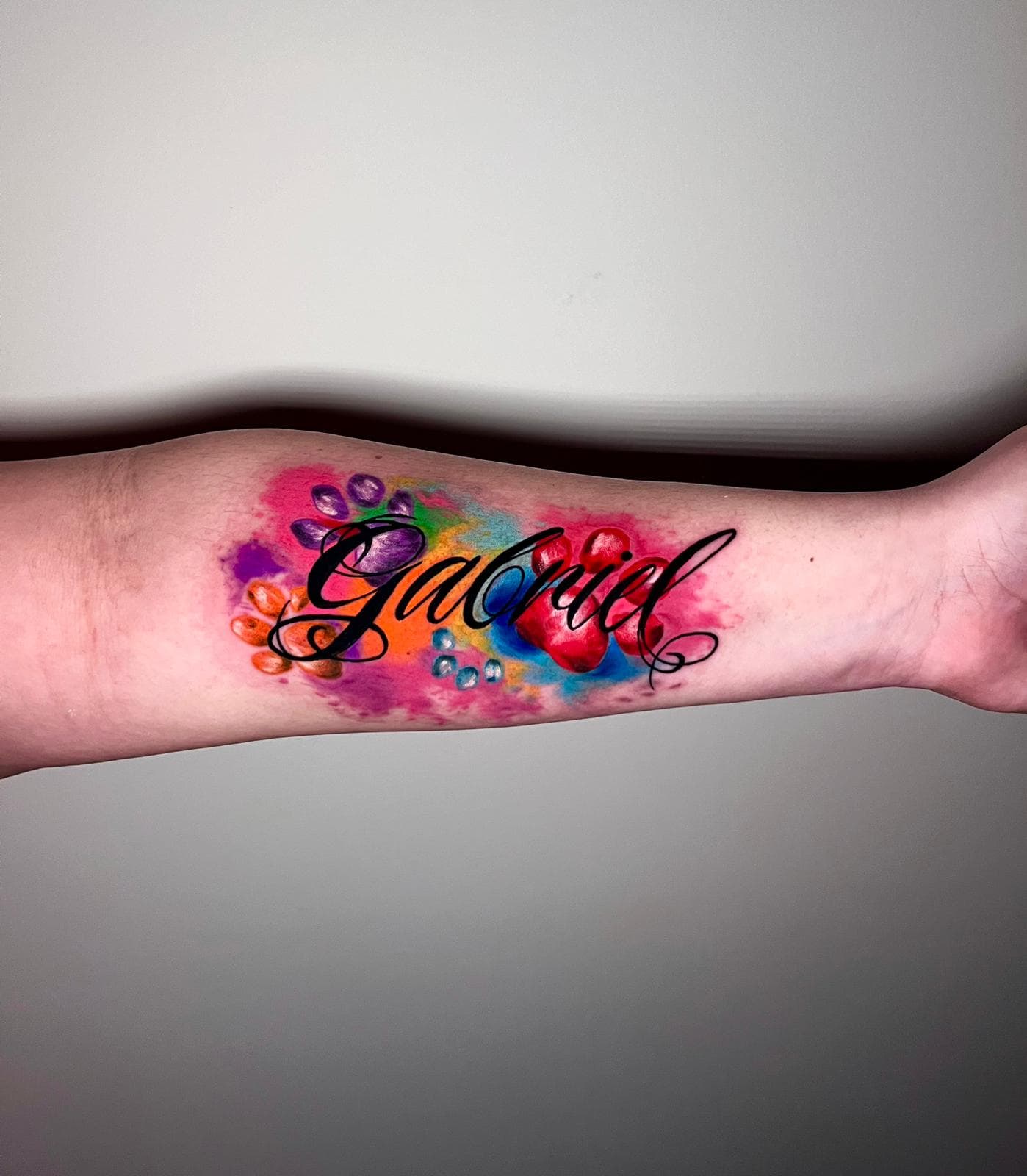 Color script name tattoo reading Gabriel on a forearm over a watercolor paw-print background